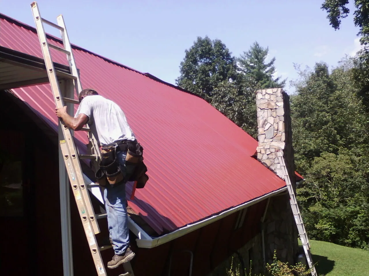Roofer on a ladder beside a red metal roof and stone chimney — Soffit & Fascia Repair by Roofing Brothers in Whitleyville